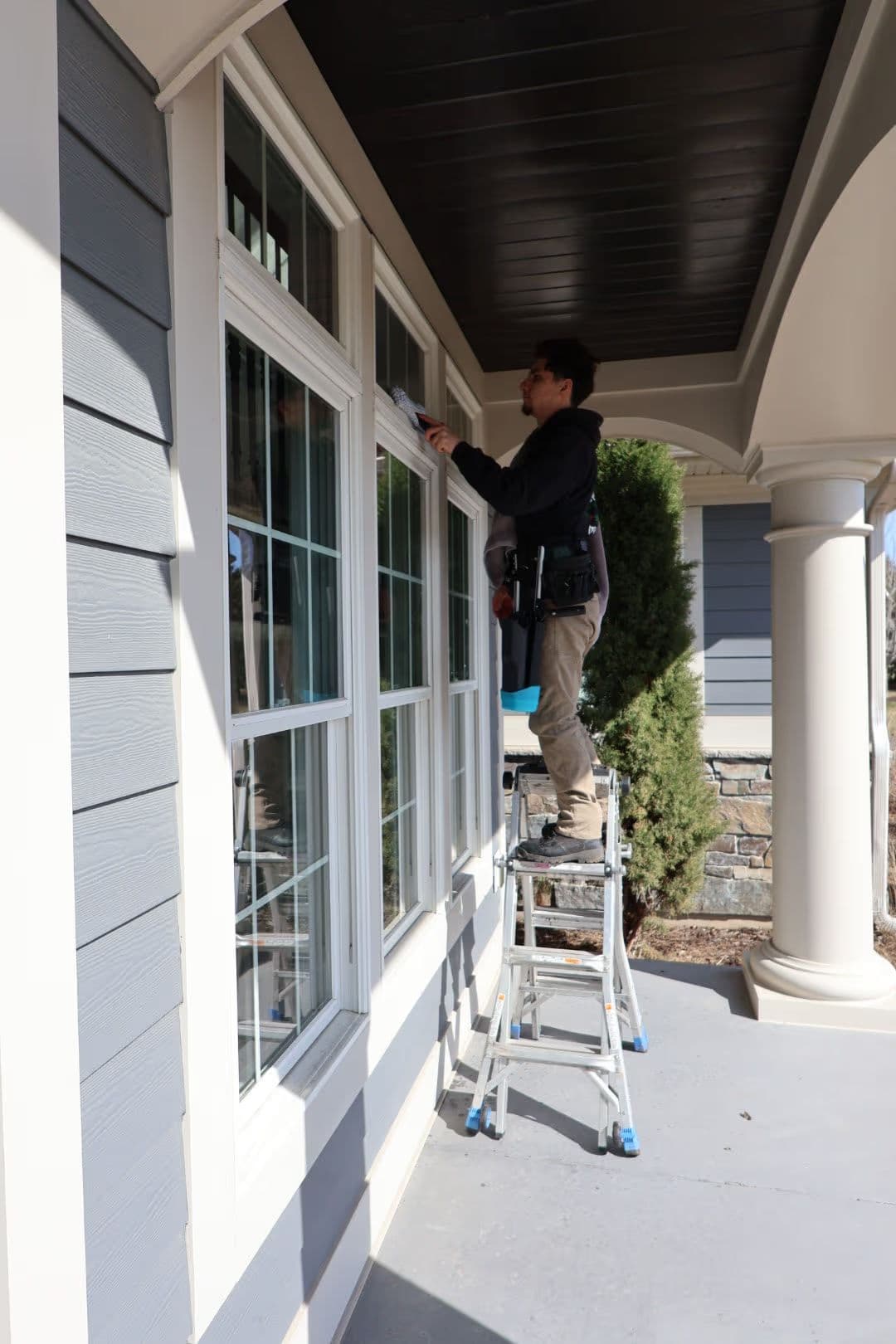 Man painting window trim while standing on a ladder outside a modern house.