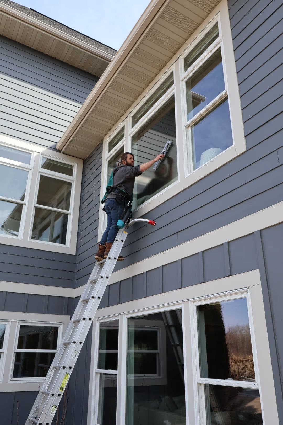 Person cleaning a large window from a ladder on a modern gray house exterior.