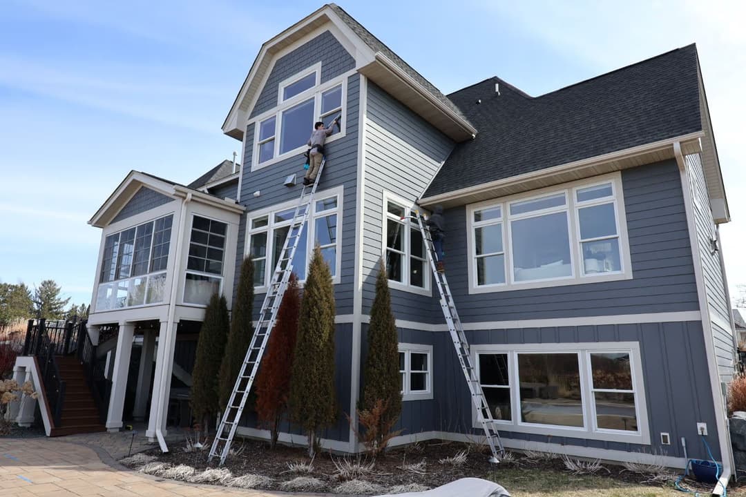 Ladders leaning against a two-story gray house with workers cleaning windows.