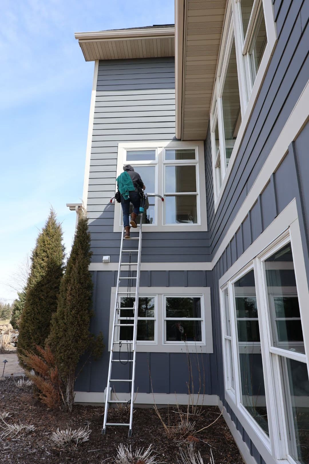 Worker on a ladder cleaning windows of a modern gray house with large windows.