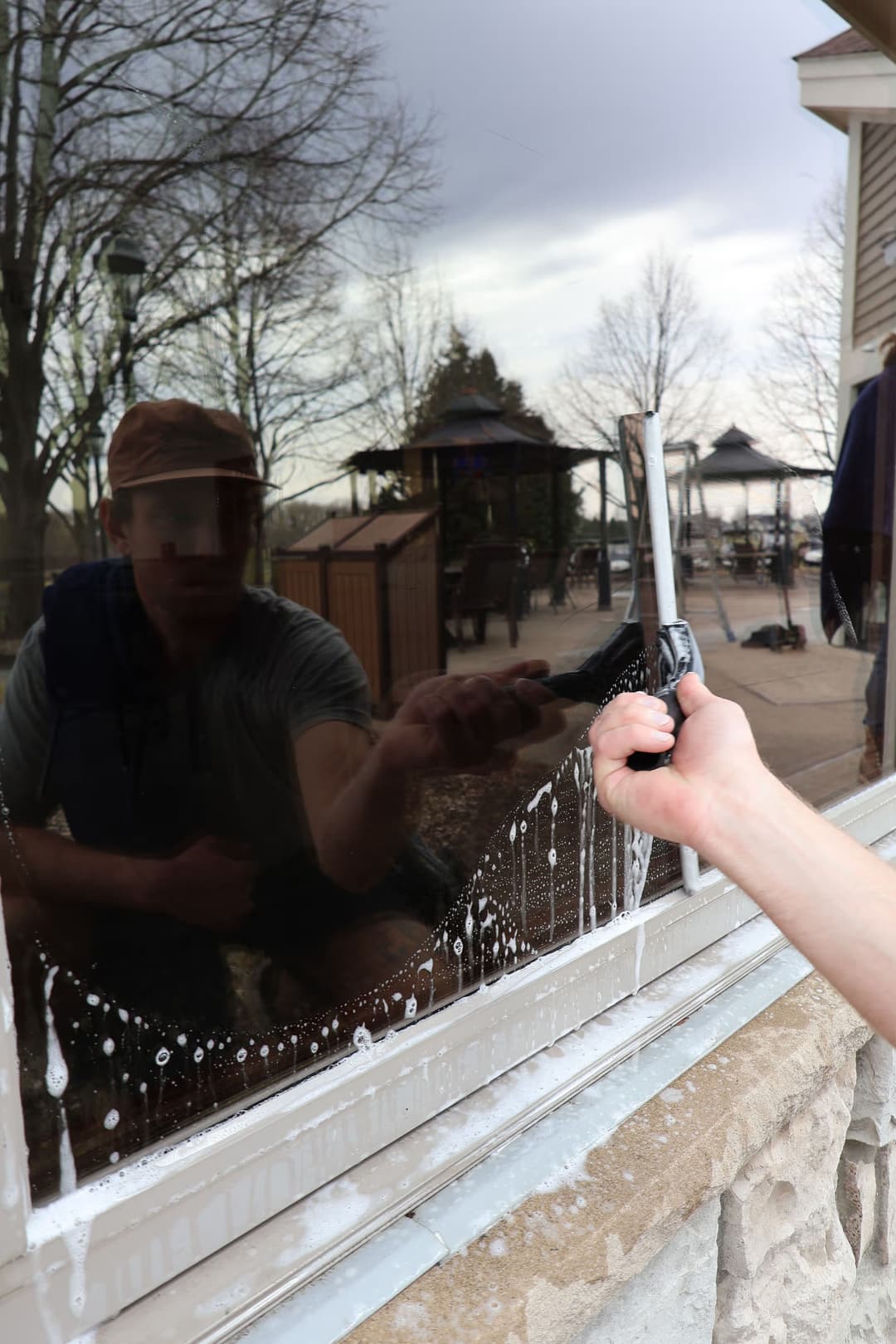 Person using a squeegee to clean a window with reflections of trees and a gazebo in the background.