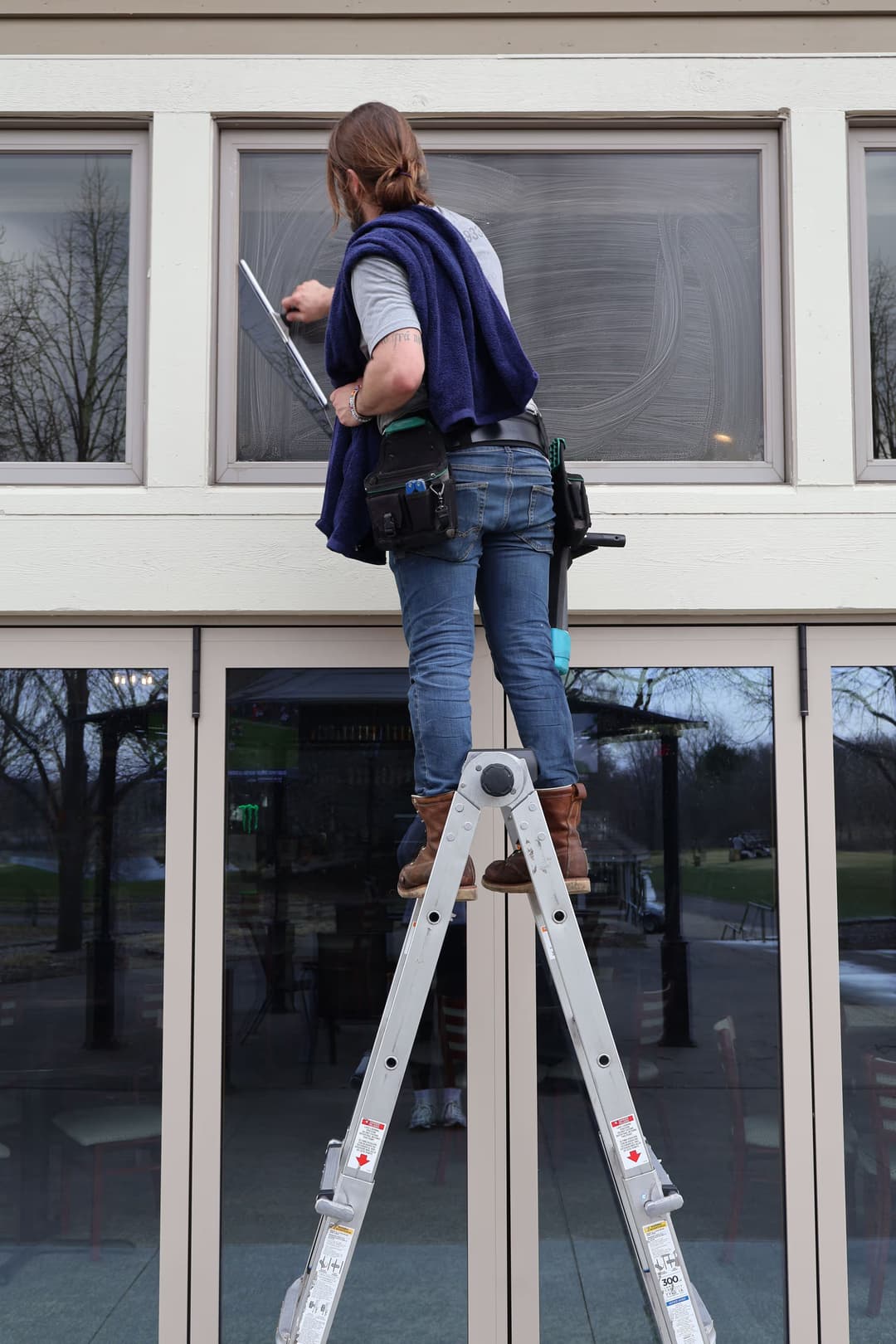 Person cleaning windows on a ladder, wearing a tool belt, with trees in the background.