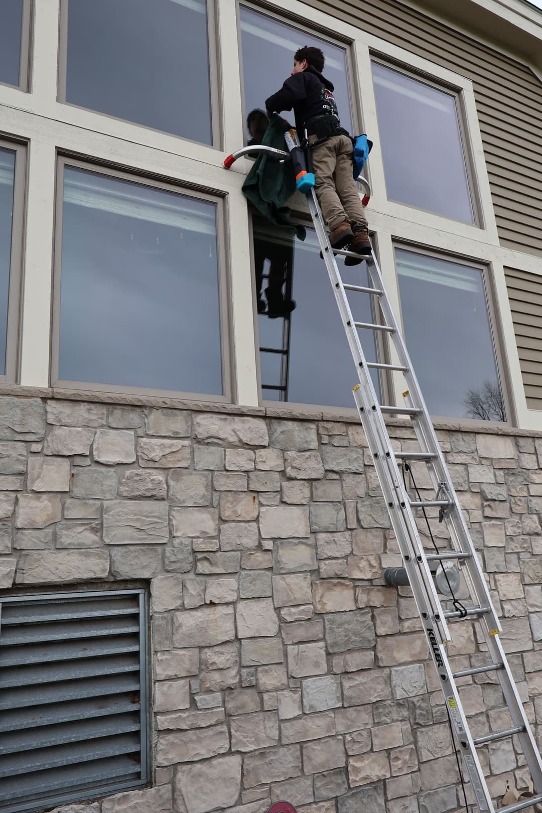 Window cleaner on ladder washing large building windows with stone facade. Safety gear visible.