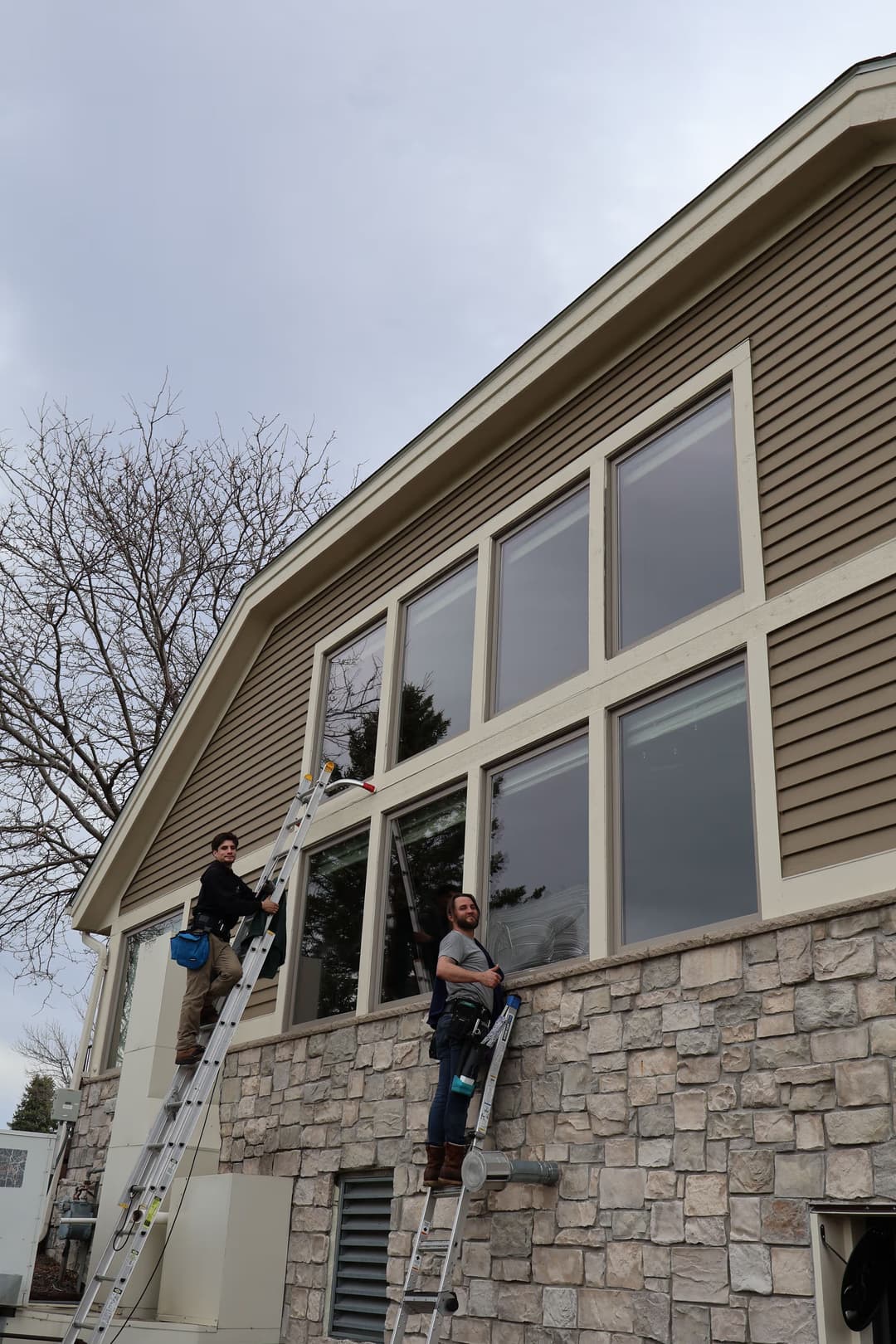 Two workers installing large windows on a house exterior with ladders, cloudy sky overhead.