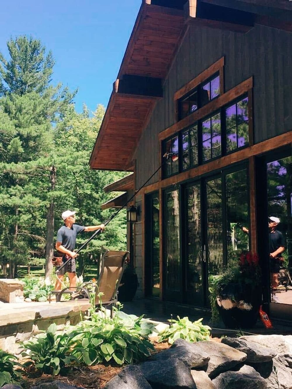 Man cleaning windows on a modern cabin surrounded by lush greenery and stone landscaping.