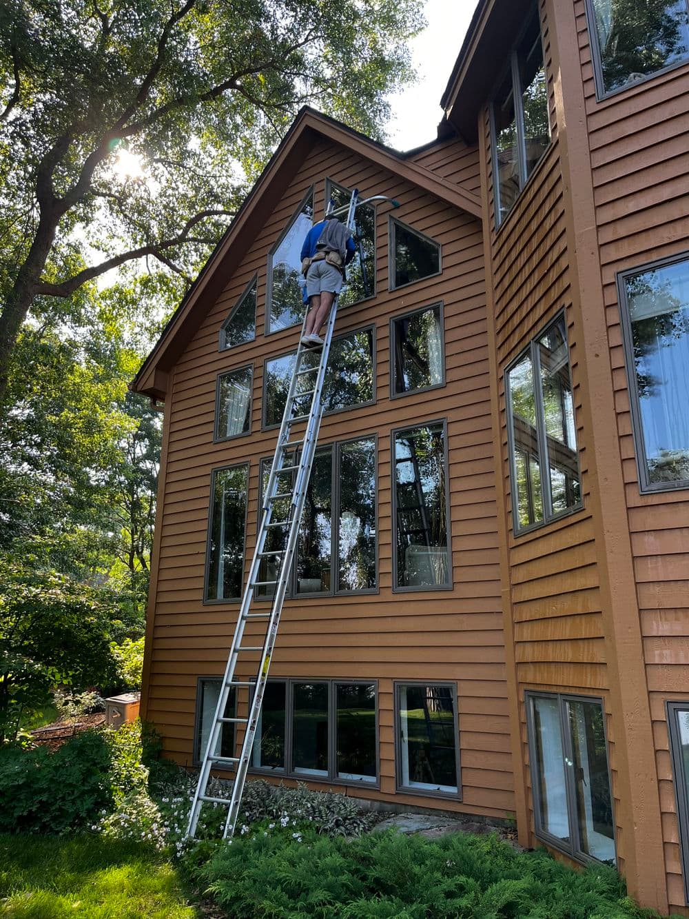 Person on ladder cleaning large windows of a modern brown house surrounded by trees.