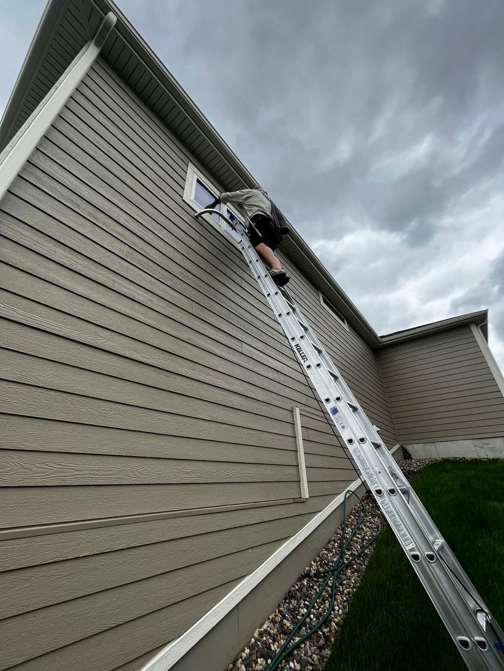 Person on ladder cleaning a window on a house with gray siding under a cloudy sky.