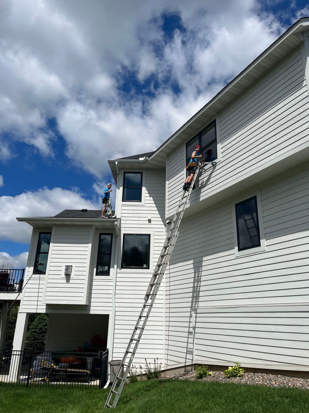 Workers using a ladder to perform maintenance on a home's upper window under a blue sky.