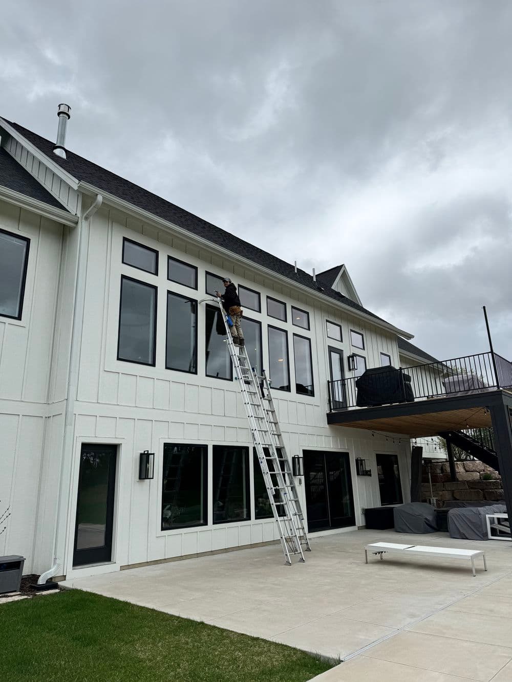 Worker on ladder cleaning large windows of a modern house under cloudy sky.