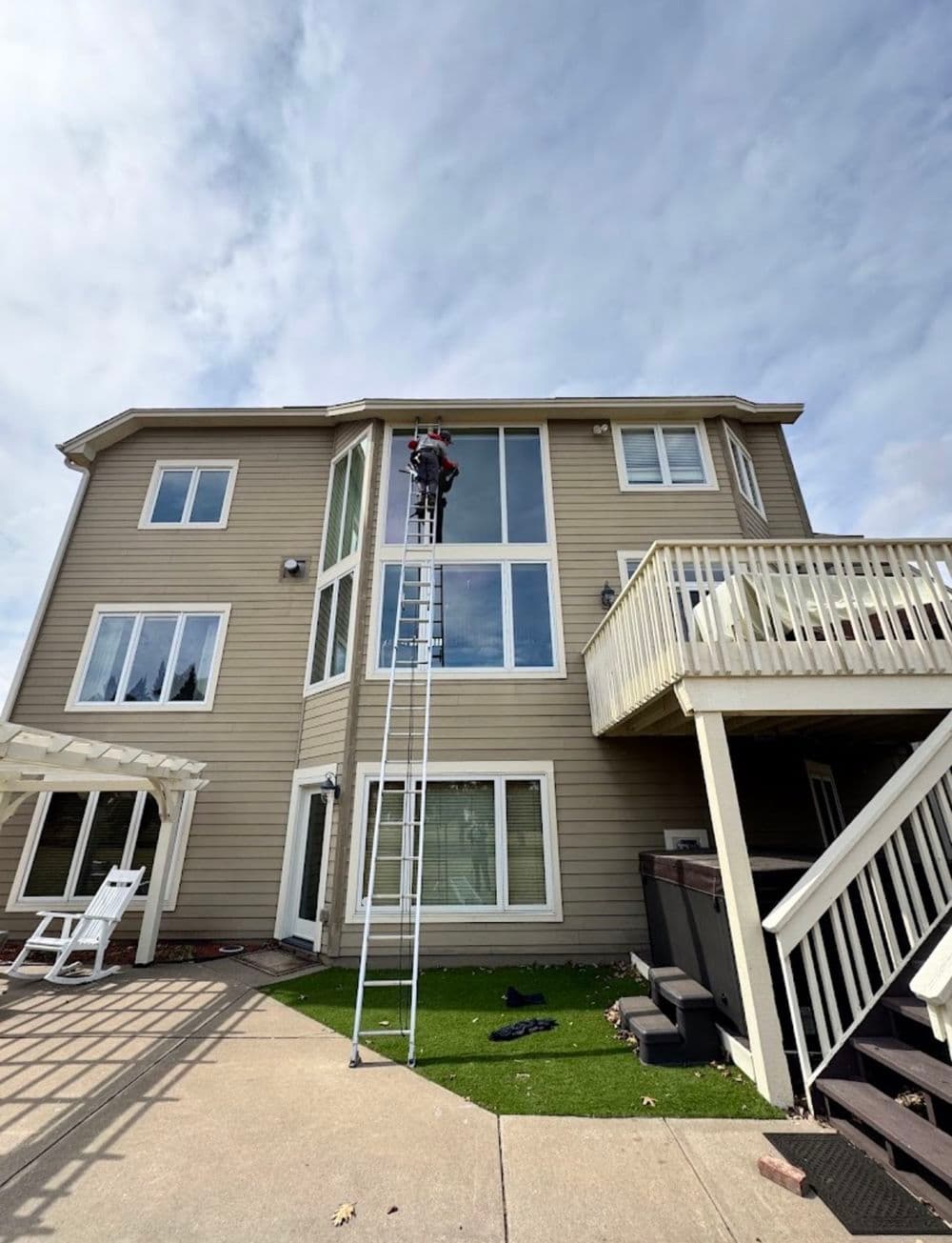Person on a ladder cleaning windows of a large two-story house with a deck.