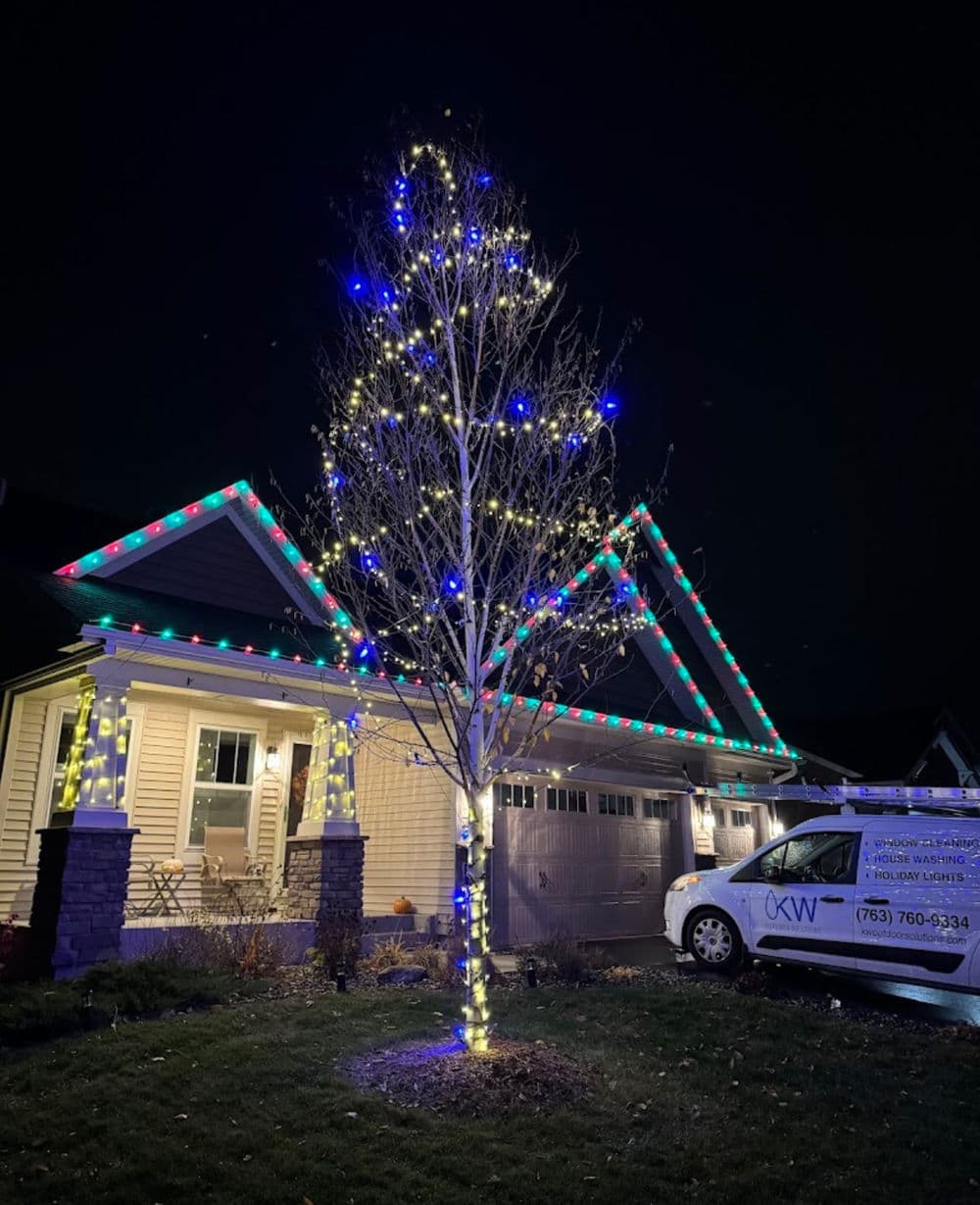 Colorful holiday lights adorn a tree and home, creating a festive nighttime scene.