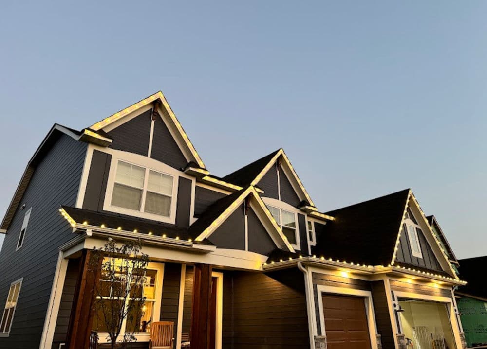 Modern house with warm LED string lights along the roofline against a clear sky.
