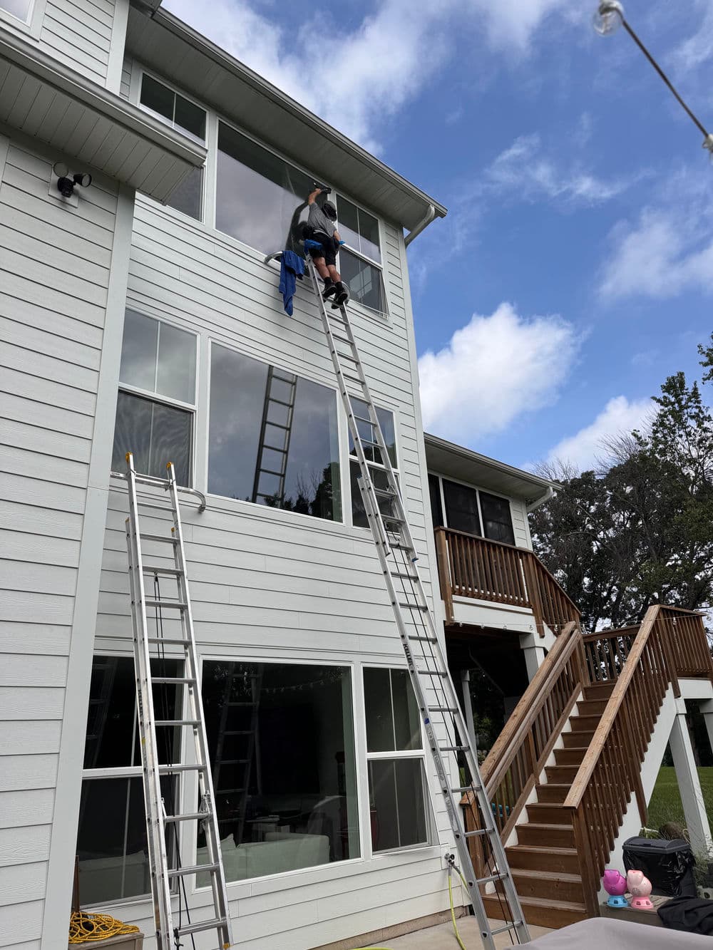 Person using a ladder to clean windows on a two-story house under a partly cloudy sky.