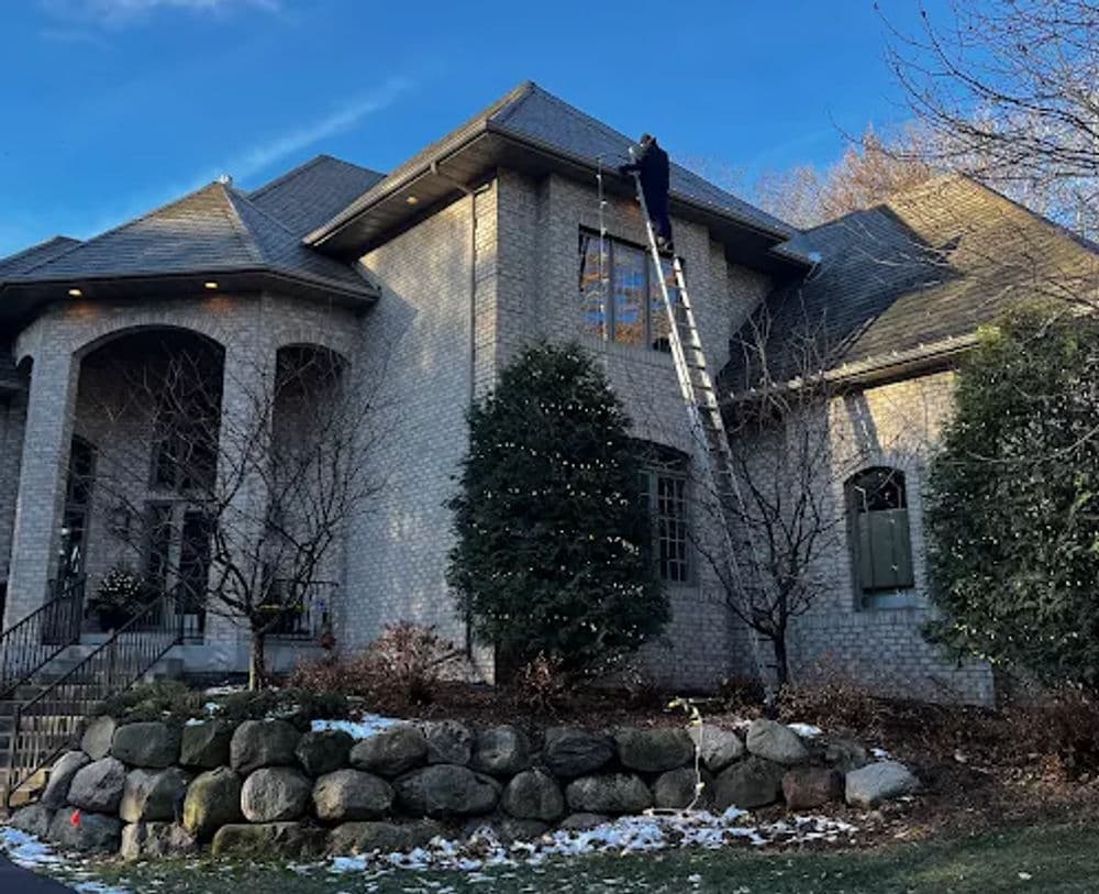 Worker cleaning windows of a large brick house using a ladder, with snowy landscape.
