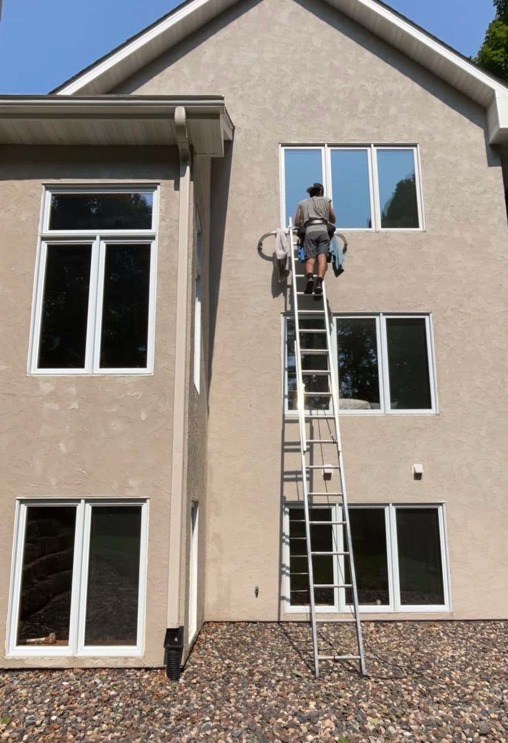 Person on ladder cleaning windows of a two-story house with gray siding.