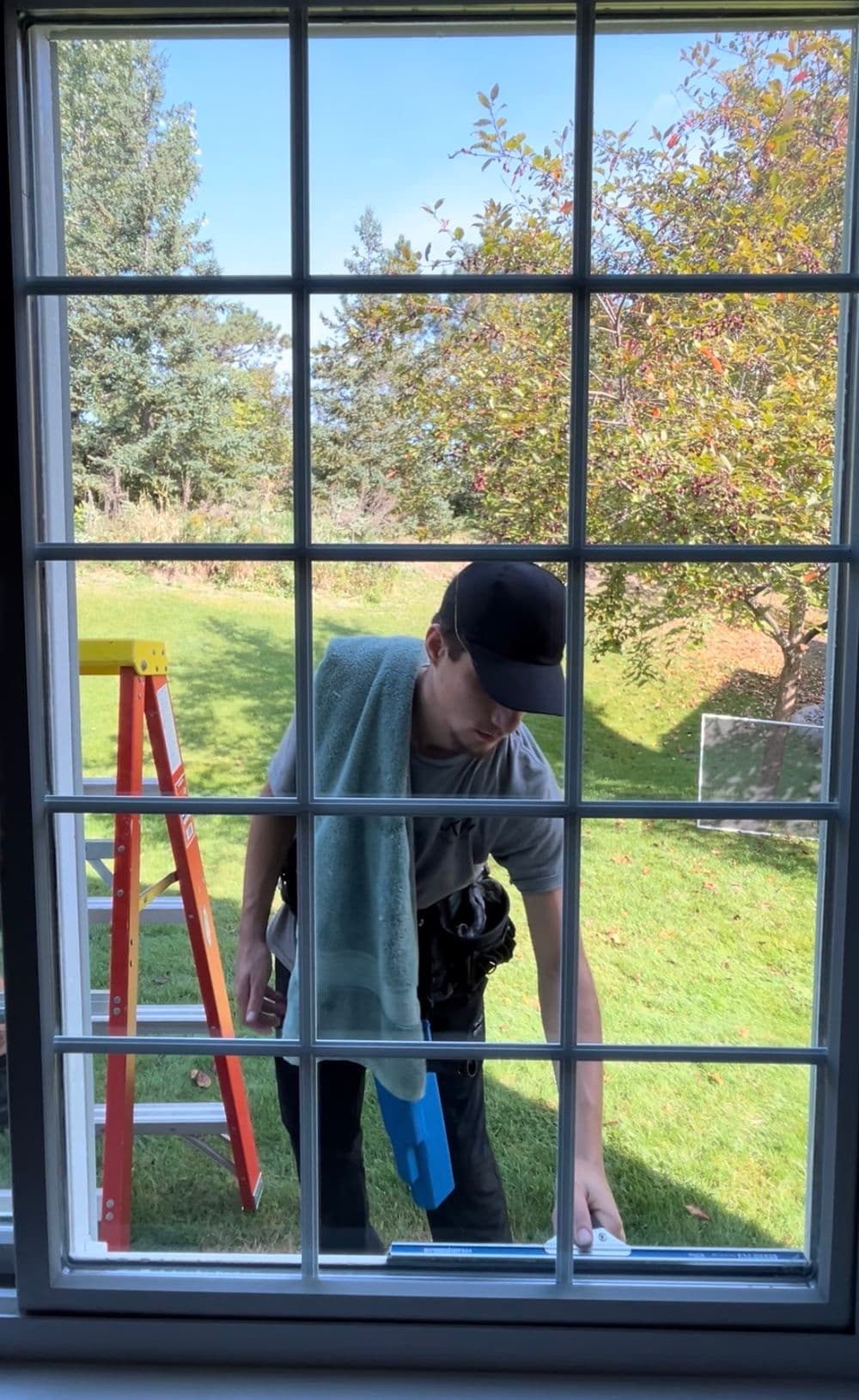 Window cleaner using a squeegee on a home, with a ladder nearby and trees in the background.