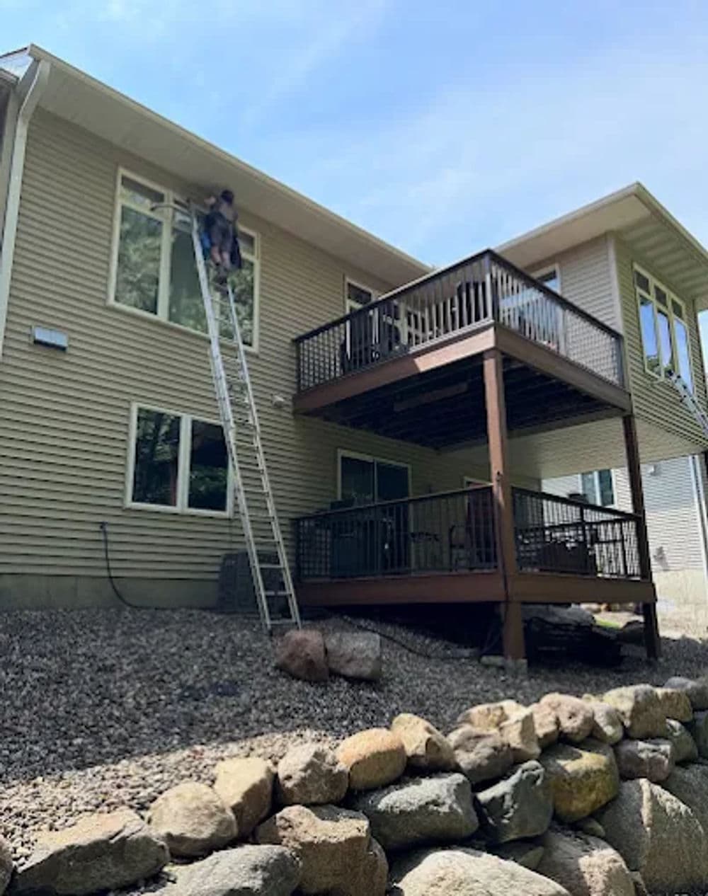 Person on a ladder cleaning windows of a two-story house with a wooden deck.