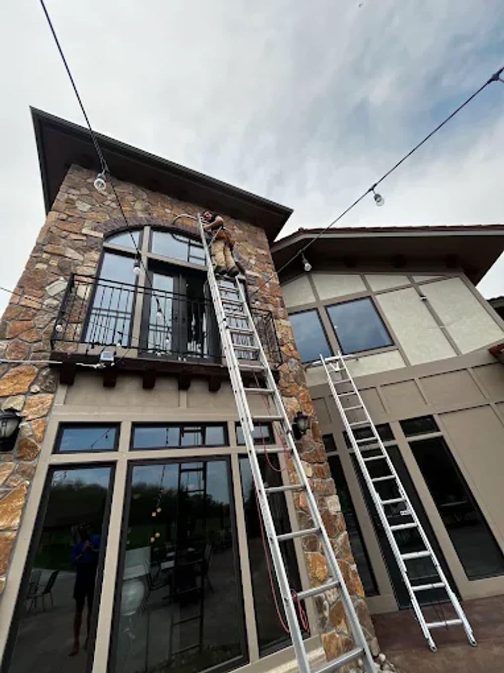 Worker using a ladder to install outdoor lights on a stone house with large windows.