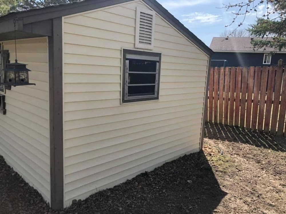 Small storage shed with beige siding and a window, set in a backyard with a wooden fence.